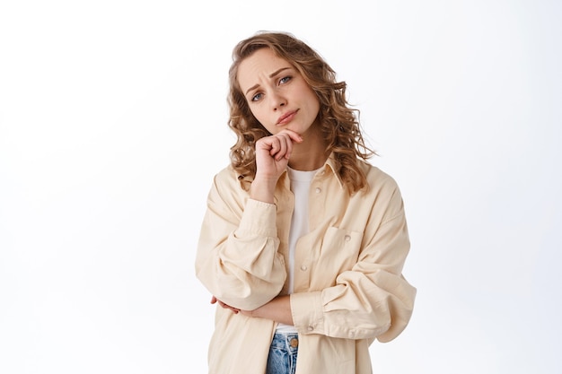 Señora indecisa mirando al frente pensativa, tomando decisiones, frunciendo el ceño y mirando pensativa, de pie con ropa moderna contra la pared blanca