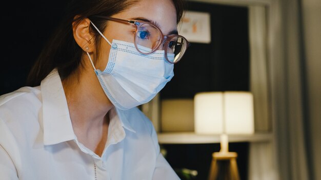 La señora independiente de Asia usa mascarilla médica, usa una computadora portátil, trabajo duro en la sala de estar de la casa.