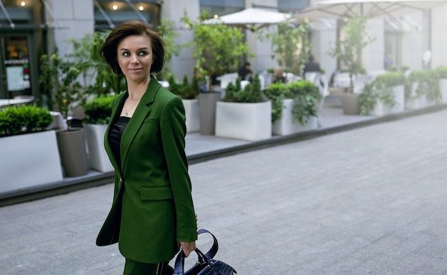 Señora independiente caminando por la calle, sosteniendo su bolso. Chaqueta verde de moda en ella. Corte de pelo corto y sexy, seguro e inteligente.