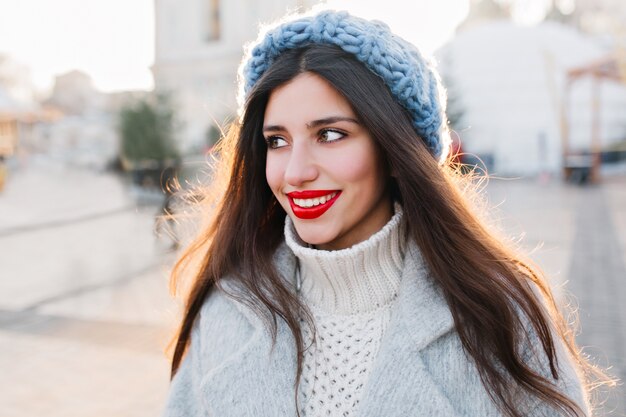 Señora inspirada de cabello oscuro con maquillaje brillante que mira hacia otro lado durante un paseo por la ciudad en invierno. Foto de primer plano de impresionante mujer morena con peinado recto soñando con algo en la calle.