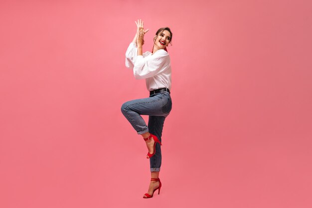 Señora en jeans y camisa blanca bailando sobre fondo rosa. Chica alegre en zapatos rojos con estilo brillante lindo sonriendo y posando para la cámara.