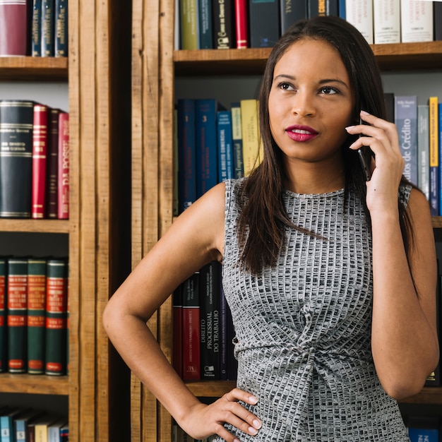 Señora joven afroamericana pensativa que habla en smartphone cerca de los libros