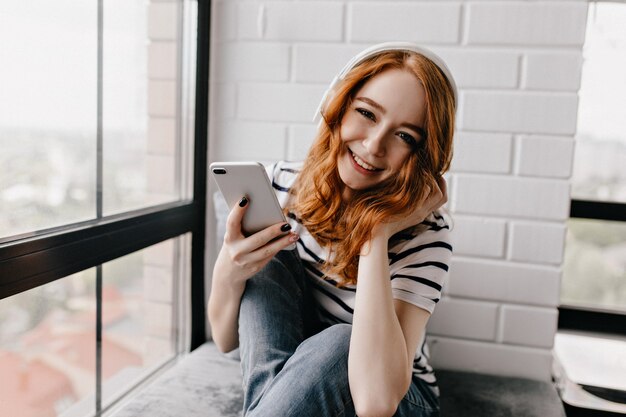 Señora joven alegre en auriculares disfrutando de fin de semana. Atractiva chica de jengibre con teléfono sentado junto a la ventana.