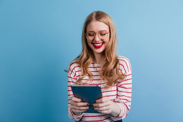 Señora joven alegre que sostiene la tableta digital con una sonrisa. Disparo de estudio de linda dama en traje casual aislado sobre fondo azul.