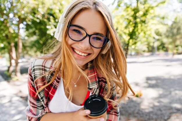 Señora joven alegre en vasos disfrutando de un café en el parque. Foto al aire libre de la feliz niña caucásica divirtiéndose al aire libre y escuchando música.