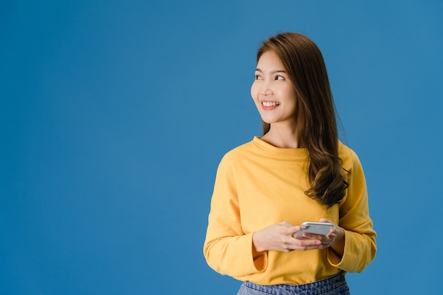 Señora joven de Asia que usa el teléfono con expresión positiva, sonríe ampliamente, vestida con ropa casual sintiendo felicidad y de pie aislado sobre fondo azul. Feliz adorable mujer alegre se regocija con el éxito.