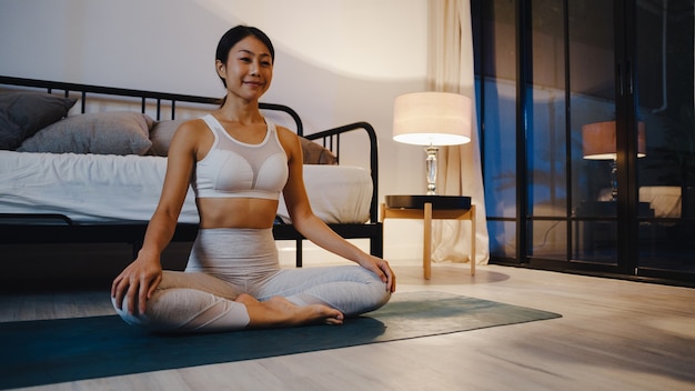 Señora joven de Asia en ropa deportiva haciendo ejercicio de yoga en la sala de estar en casa por la noche.