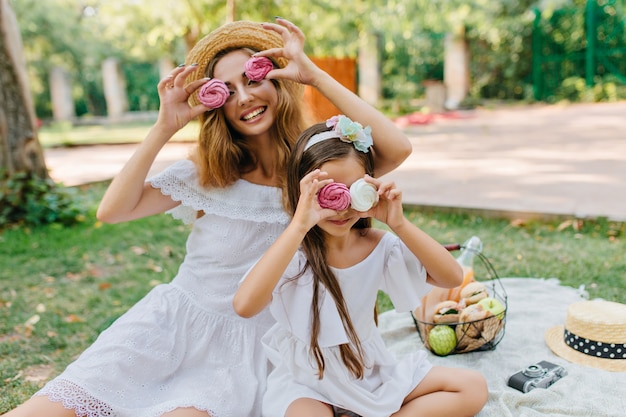 Señora joven atractiva con sombrero de paja retro bromeando con su hija y jugando con galletas de colores. Dos lindas hermanas haciendo un picnic en el parque de verano y riendo.