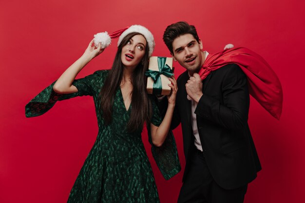Señora joven atractiva en vestido verde y gorro de Papá Noel con caja de regalo y posando con una hermosa morena con traje negro