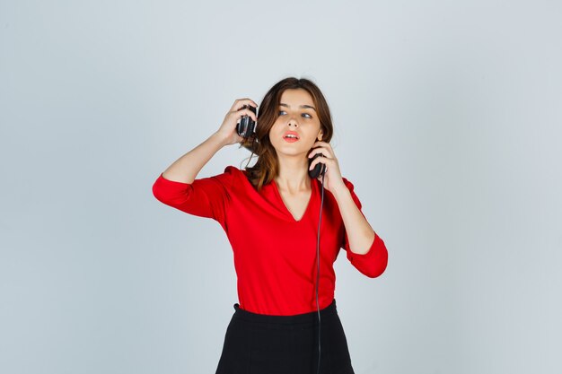 Señora joven con auriculares en blusa roja, falda posando mientras está de pie y luciendo bonita