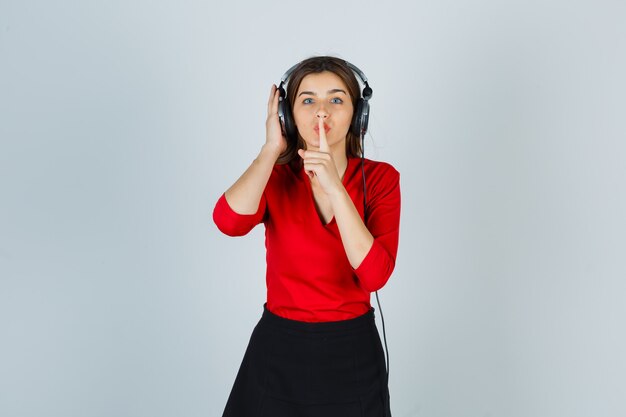Señora joven con auriculares escuchando música mientras muestra gesto de silencio en blusa roja