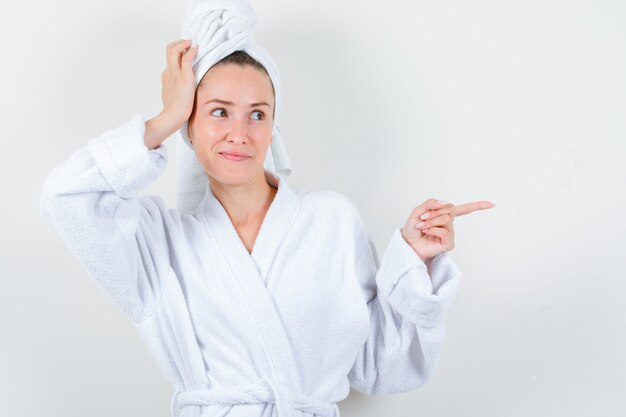 Señora joven en bata de baño blanca, toalla sosteniendo la mano en la cabeza mientras apunta hacia el lado derecho y parece complacida, vista frontal.