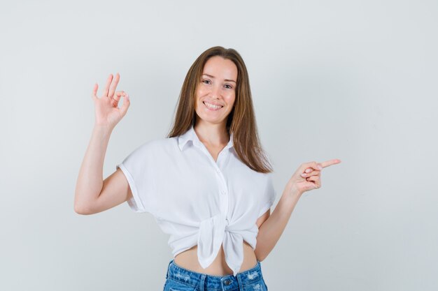 Señora joven en blusa blanca que muestra un gesto aceptable mientras apunta a un lado y mira positiva, vista frontal.