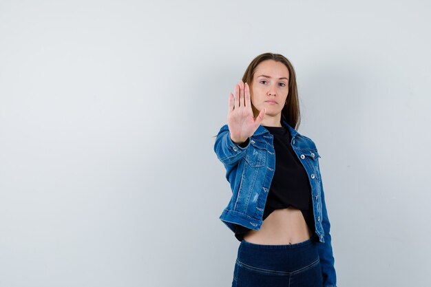 Señora joven en blusa, chaqueta, jeans mostrando gesto de parada y mirando confiado, vista frontal.