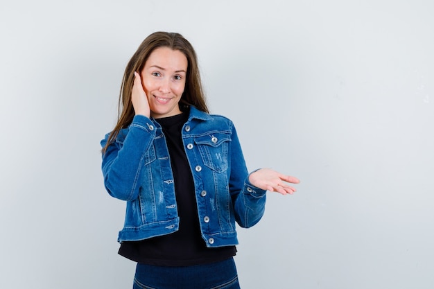 Señora joven en blusa, chaqueta manteniendo la palma abierta y mirando alegre, vista frontal.