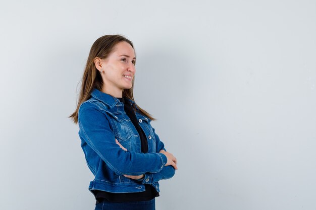 Señora joven en blusa, chaqueta de pie con los brazos cruzados y mirando soñadora, vista frontal.