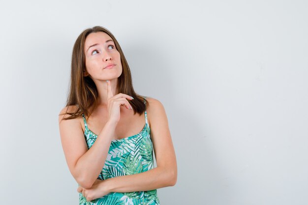 Señora joven en blusa de pie en pose de pensamiento y mirando pensativo, vista frontal.