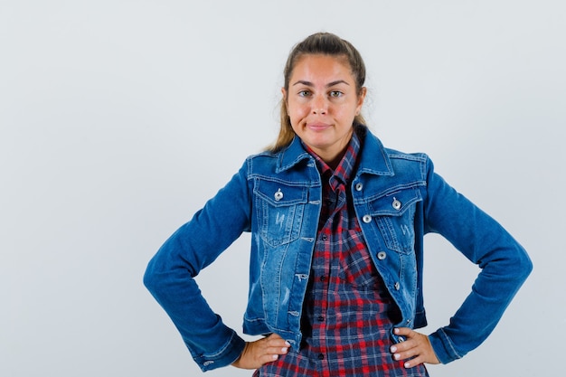 Señora joven en camisa, chaqueta cogidos de la mano en la cintura y mirando confiado, vista frontal.