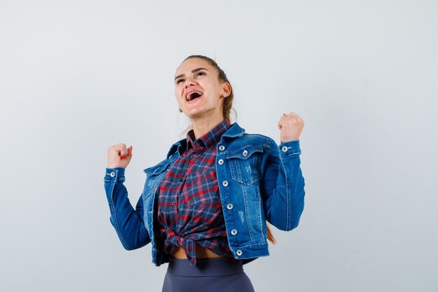 Señora joven en camisa, chaqueta que muestra el gesto del ganador mientras grita y se ve feliz, vista frontal.