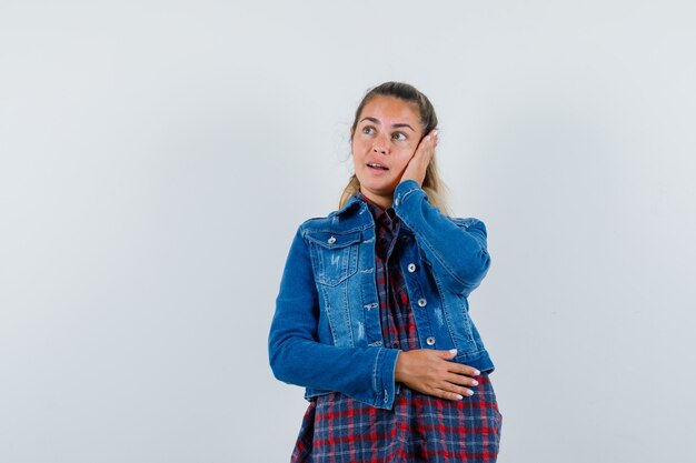 Señora joven en camisa, chaqueta sosteniendo la mano en la mejilla y mirando soñadora, vista frontal.