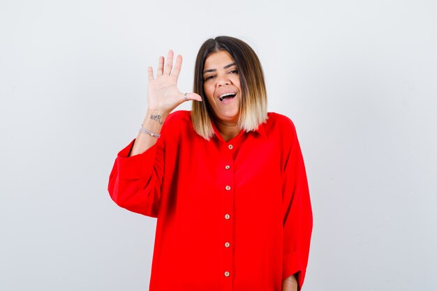 Señora joven en camisa roja de gran tamaño agitando la mano para saludar y mirar alegre, vista frontal.