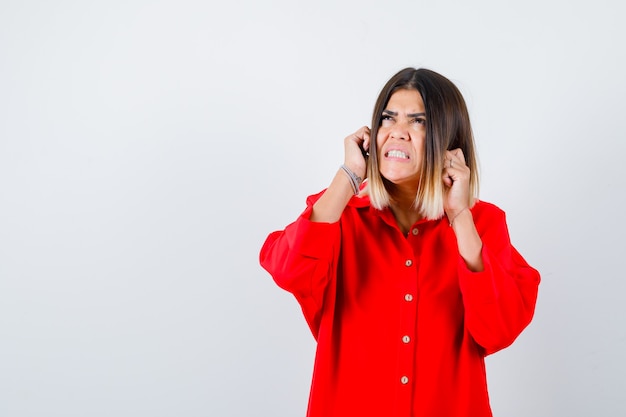 Señora joven en camisa roja de gran tamaño sosteniendo los puños cerca de la cara y mirando molesto, vista frontal.