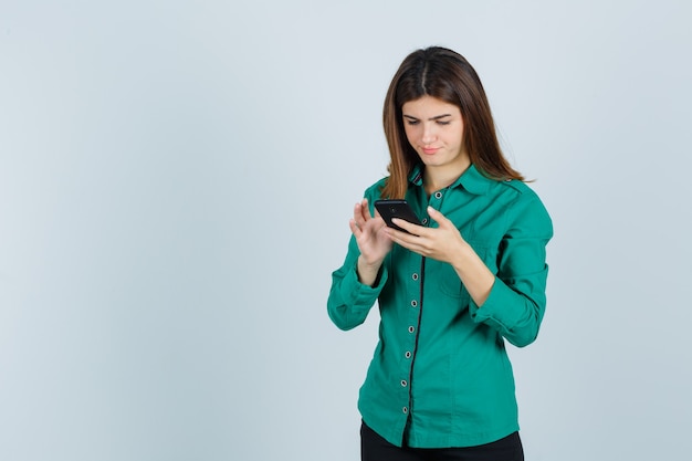Señora joven en camisa verde escribiendo en el teléfono móvil y mirando ocupado, vista frontal.