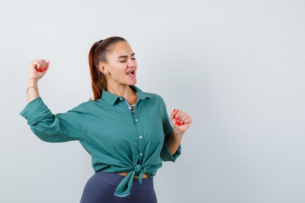 Señora joven en camisa verde que muestra el gesto del ganador y mirando dichoso, vista frontal.