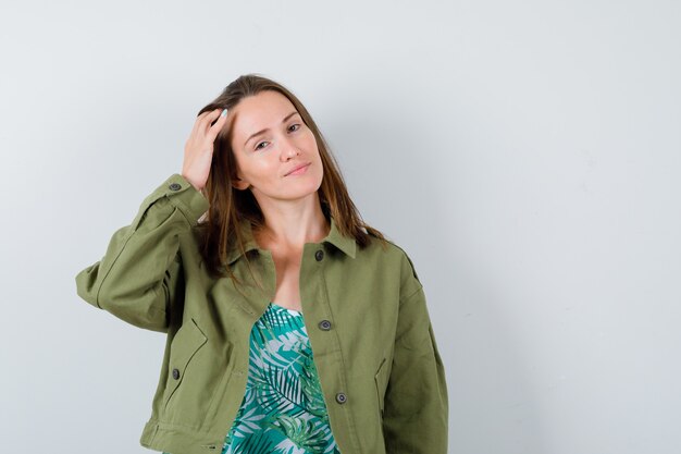 Señora joven en chaqueta verde posando con la mano en la cabeza y mirando seductor, vista frontal.