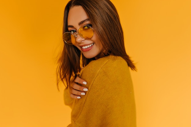Foto gratuita señora joven complacida con cabello castaño brillante posando en la pared amarilla. encantadora chica caucásica con gafas de sol mirando por encima del hombro.