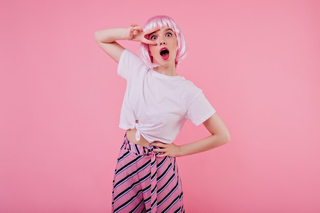 Señora joven glamorosa en traje de verano de moda lleva peluca rosa. Foto interior de modelo femenino positivo en camiseta blanca relajándose durante la sesión de fotos