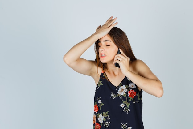 Foto gratuita señora joven hablando por teléfono inteligente, sosteniendo la mano en la cabeza en la blusa y mirando nerviosa, vista frontal.