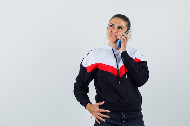 Señora joven hablando por teléfono en sudadera colorida y mirando pensativo, vista frontal.