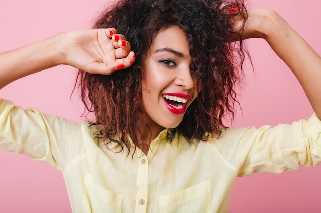 Señora joven inspirada con manicura roja posando con las manos arriba y una sonrisa sincera. Elegante mujer mulata con peinado corto que expresa felicidad.