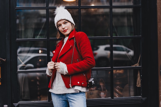 Señora joven con lápiz labial rojo y ojos marrones vestida con abrigo brillante y sombrero blanco posando con cámara retro contra la ventana.