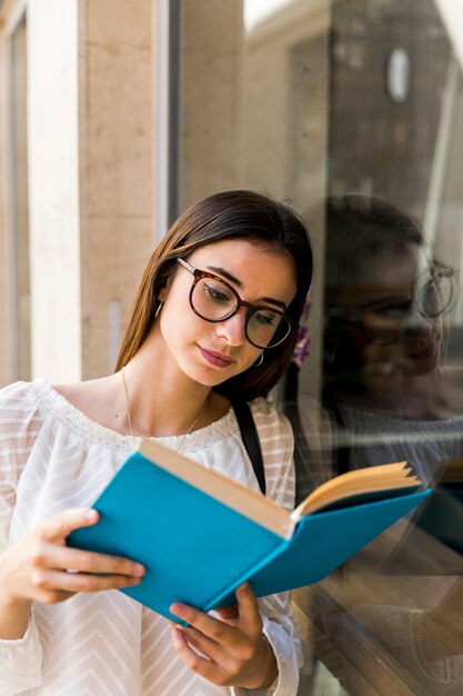 Señora joven con el libro de lectura de los vidrios cerca de la ventana