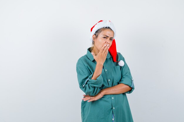 Señora joven con la mano en la boca con sombrero de Navidad, camisa y mirando pensativo, vista frontal.