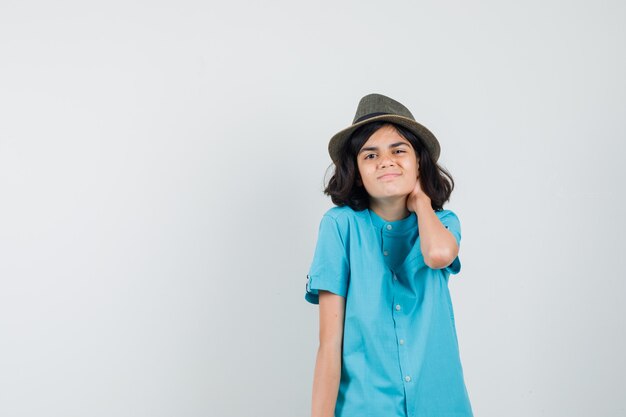 Señora joven con la mano en el cuello con camisa azul, sombrero y aspecto cansado.