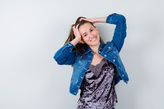 Señora joven con las manos en la cabeza en blusa, chaqueta vaquera y mirando feliz. vista frontal.