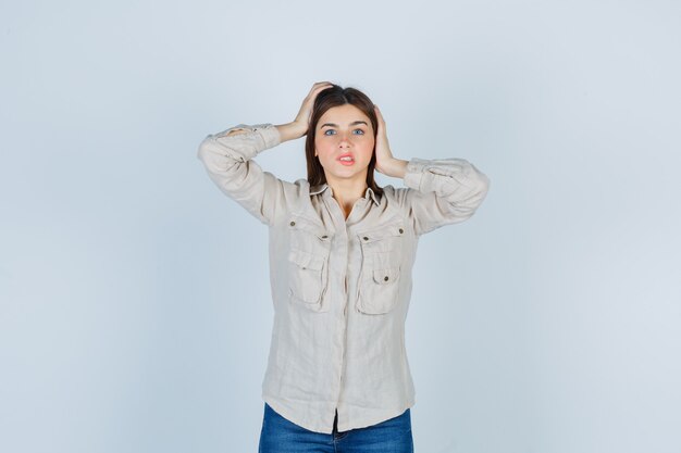 Señora joven con las manos en la cabeza en casual, jeans y mirando preocupado, vista frontal.