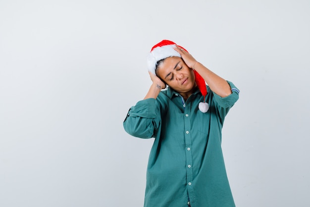 Señora joven con las manos en la cabeza con sombrero de navidad, camisa y mirando relajado, vista frontal.