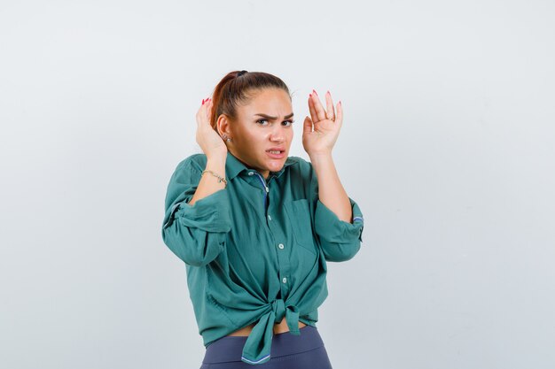 Señora joven con las manos cerca de la cabeza en camisa, pantalones y mirando ansiosa, vista frontal.