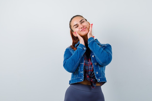 Señora joven con las manos en las mejillas en camisa a cuadros, chaqueta de mezclilla y mirando pacífica, vista frontal.