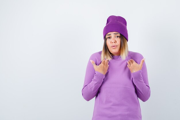 Señora joven con las manos en el pecho en suéter morado, gorro y mirando ansiosa. vista frontal.