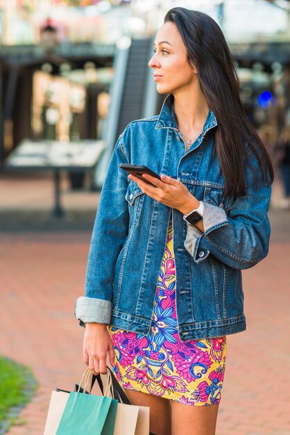 Señora joven con los paquetes de las compras que sostienen el teléfono móvil en centro comercial