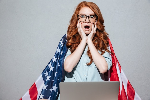 Señora joven pelirroja con bandera de Estados Unidos usando la computadora portátil