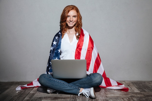 Señora joven pelirroja con bandera de Estados Unidos usando la computadora portátil