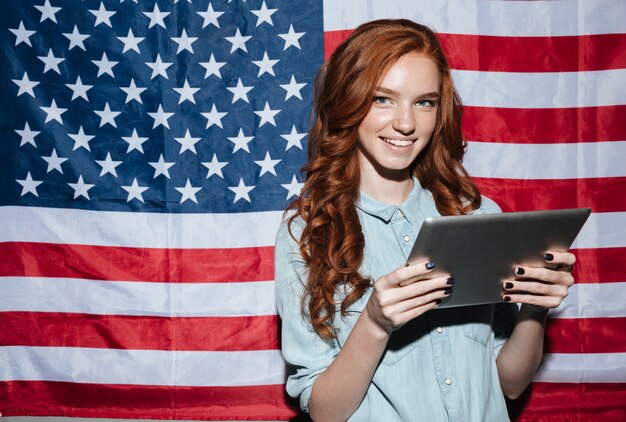 Señora joven pelirroja de pie sobre la bandera de Estados Unidos usando tableta