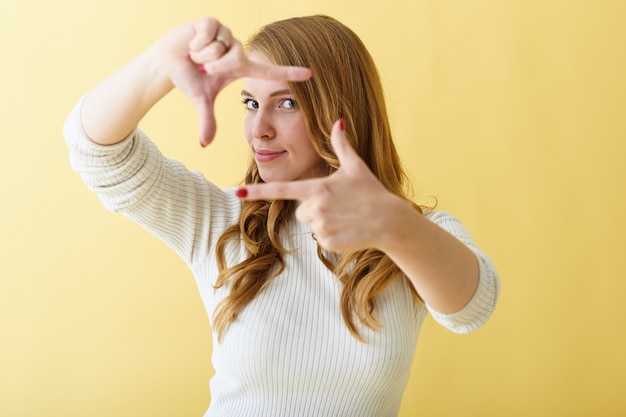 Foto gratuita señora joven positiva de moda con uñas rojas cuidadas gesticulando, haciendo el marco de la cámara con sus dedos, posando aislado contra el fondo de la pared del espacio de copia amarilla en blanco para su texto o anuncio