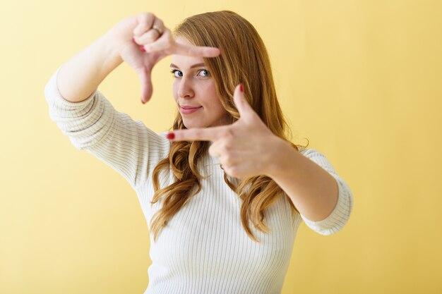 Señora joven positiva de moda con uñas rojas cuidadas gesticulando, haciendo el marco de la cámara con sus dedos, posando aislado contra el fondo de la pared del espacio de copia amarilla en blanco para su texto o anuncio